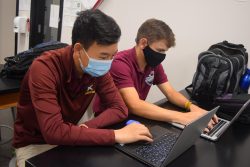 Two high school students, seated next to each other, work on laptops.